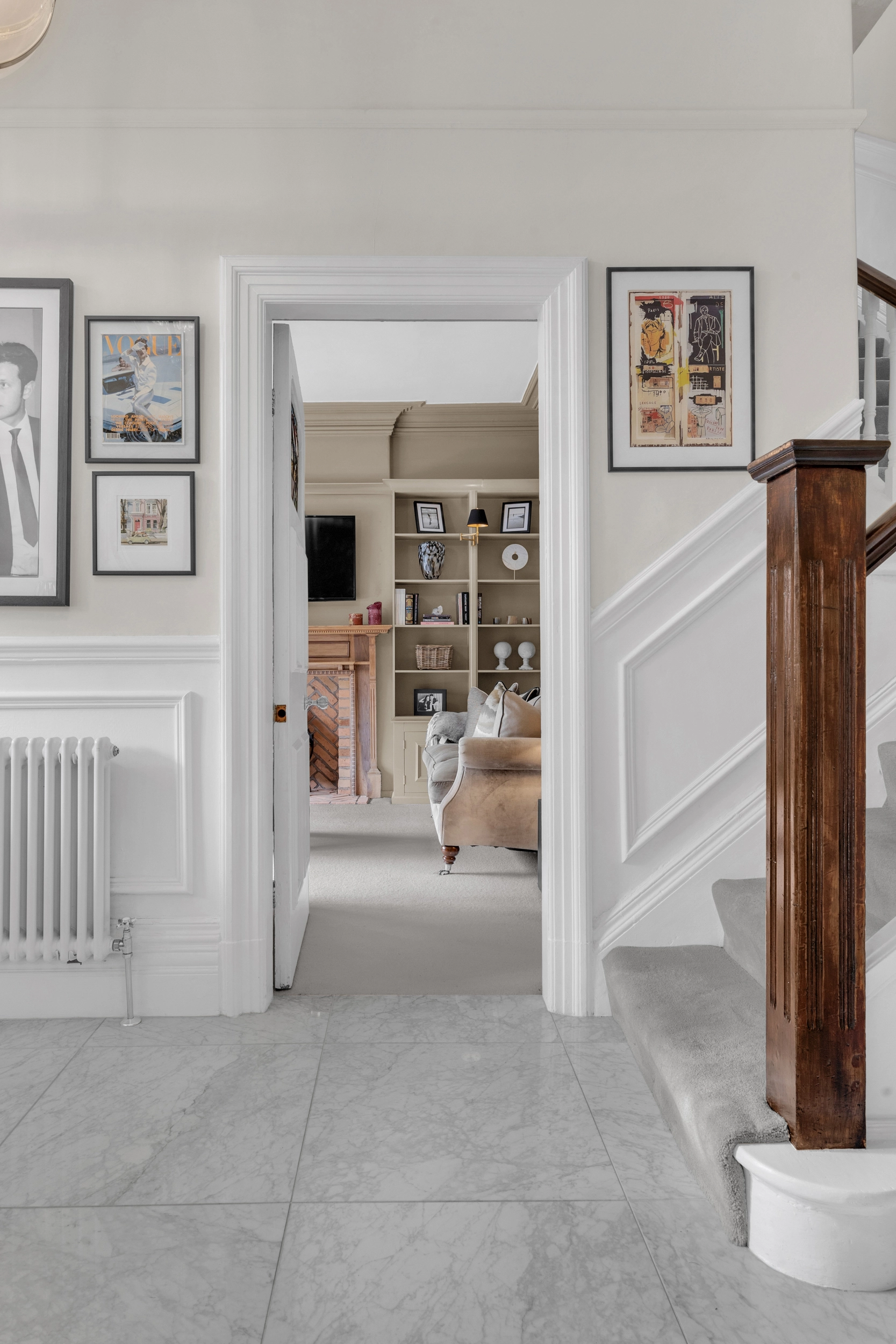 Hallway with white walls and grey tiled floor, leading through open doorway to bedroom with built-in shelving and beige armchair