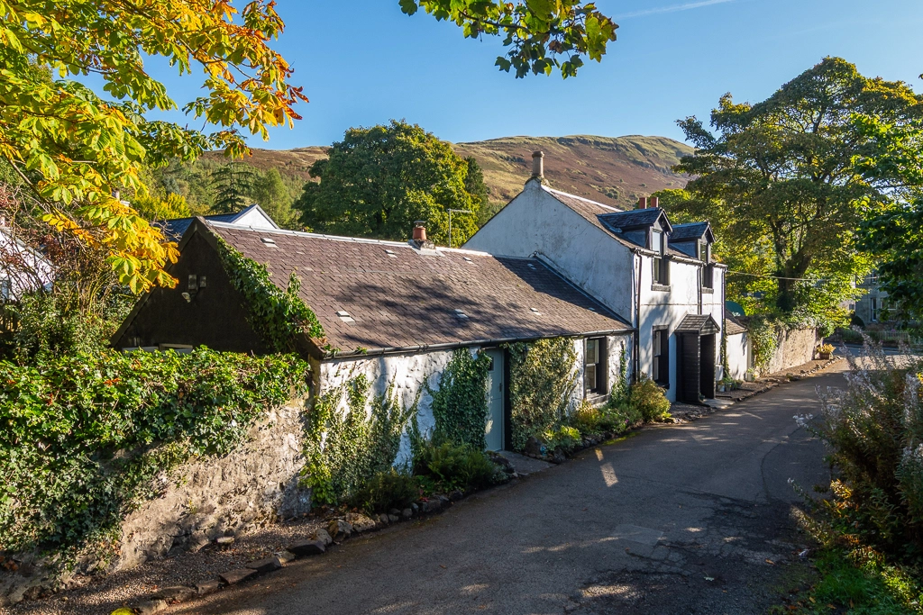 White-rendered cottage with slate roof and ivy-covered walls, set on a gravel driveway with rolling hills in the background