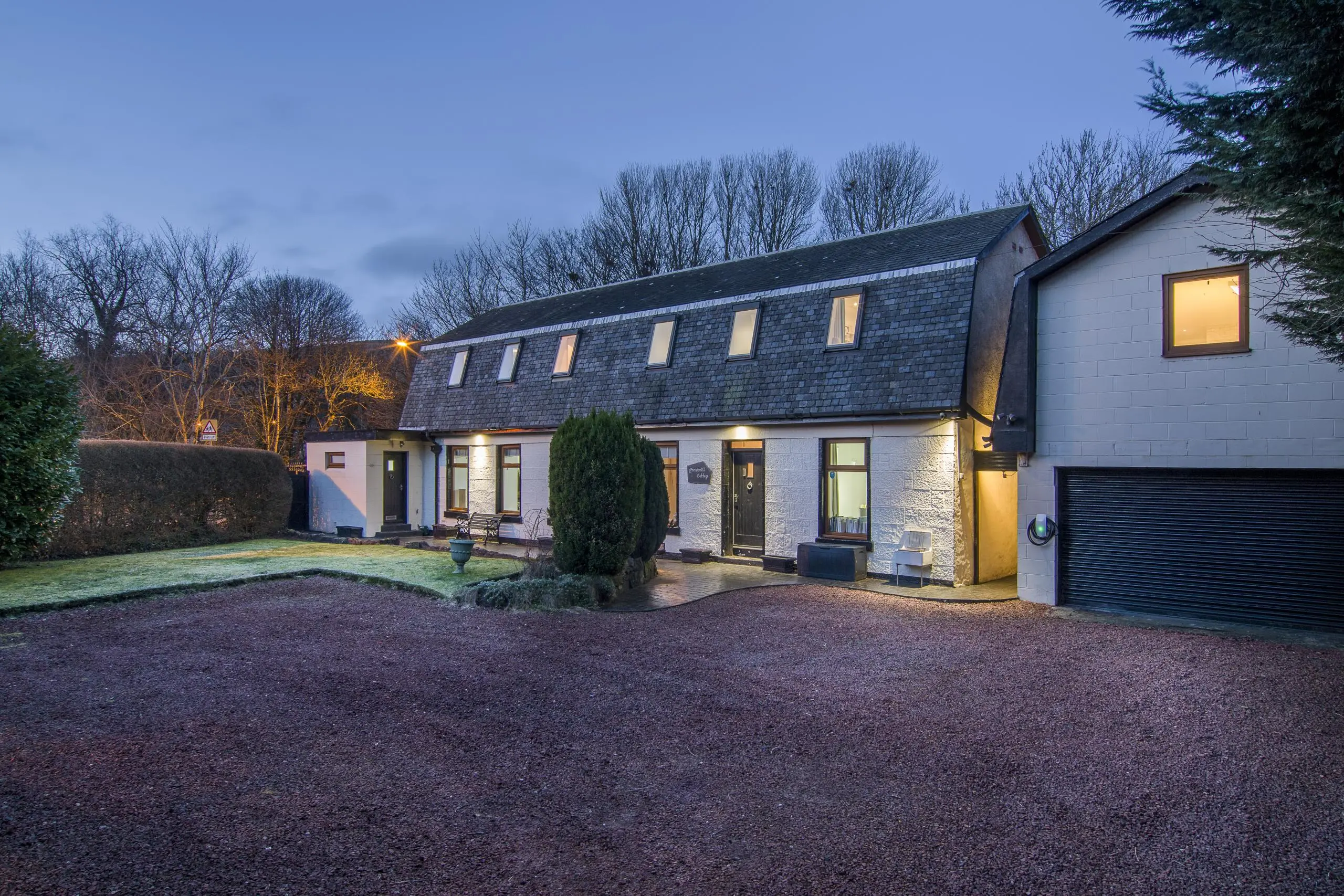 Modern white-rendered house with slate roof and integrated garage, lit at dusk with gravel drive and landscaped garden