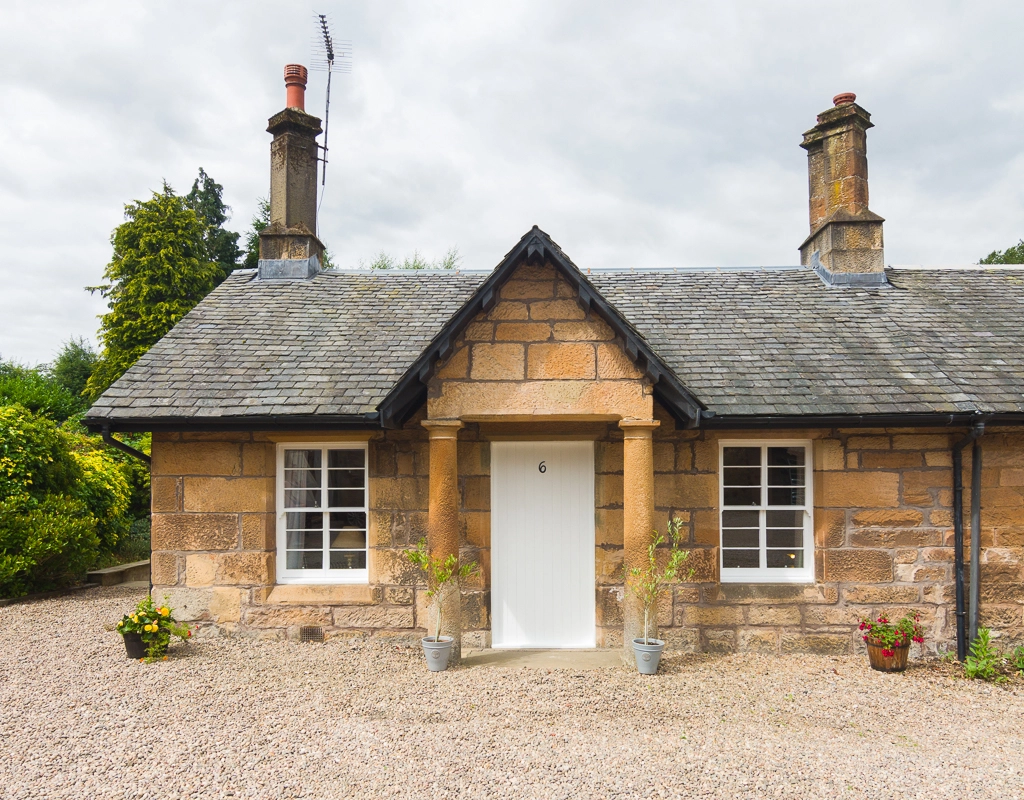 Stone cottage with white door, two windows, slate roof and two chimneys, gravel driveway with potted plants