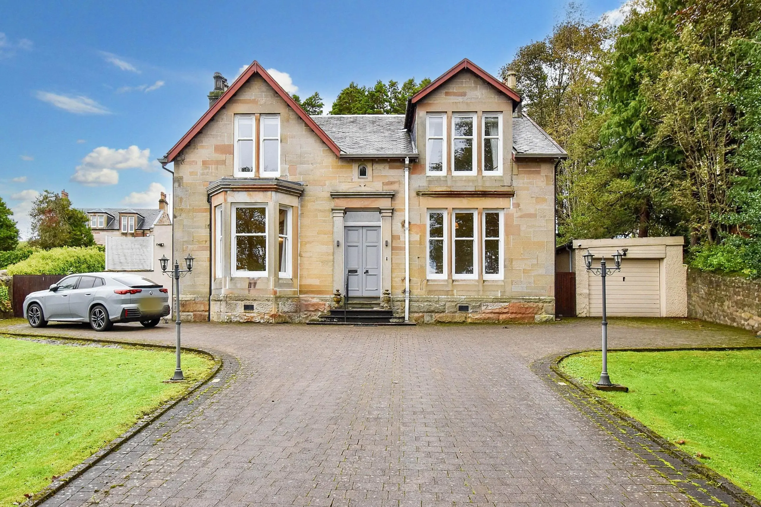 Stone-built detached house with red roof, white-framed windows, grey front door, driveway with parked silver car, and manicured lawn
