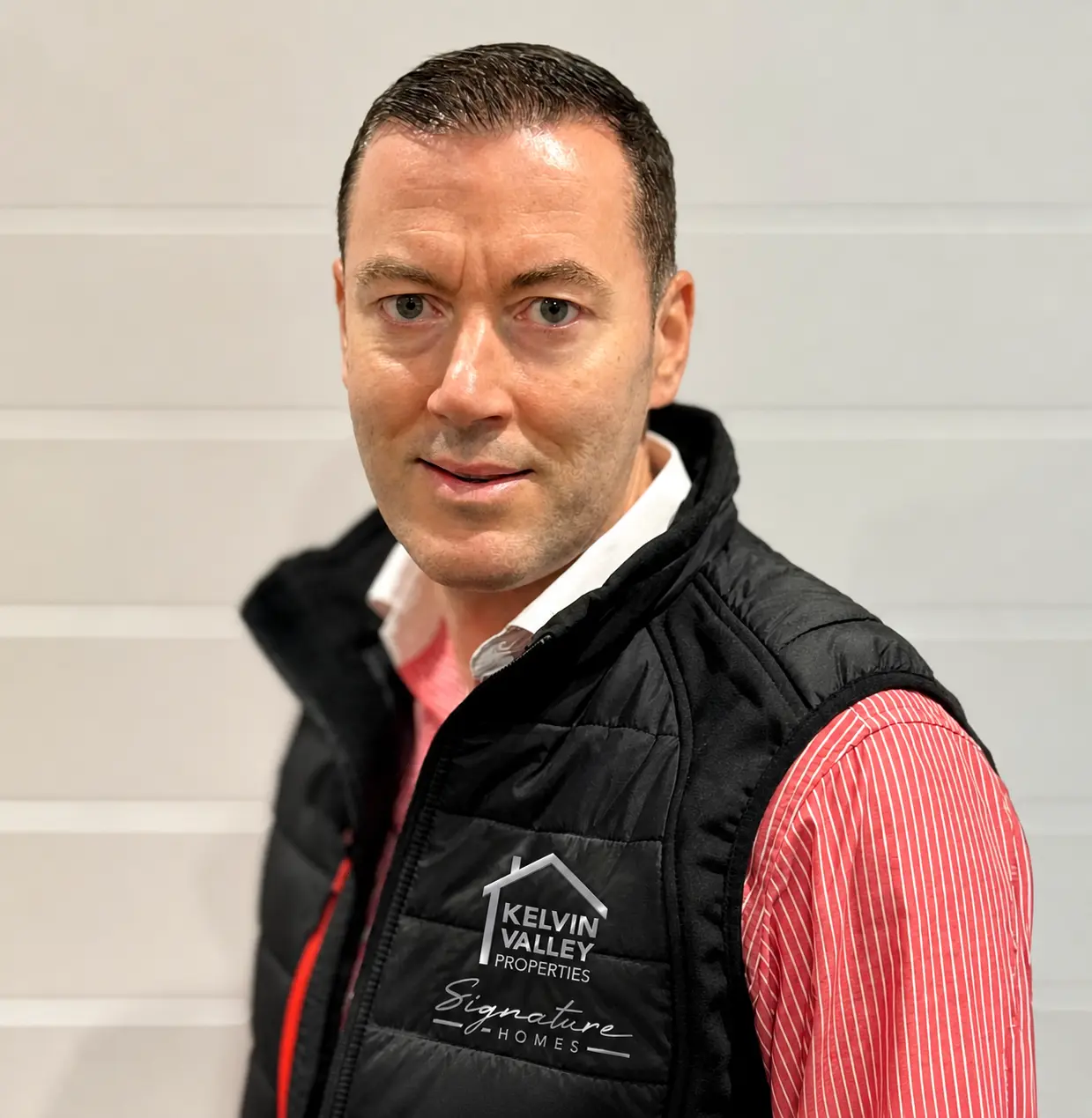 Man wearing black quilted gilet with "Kelvin Valley Properties Signature Homes" logo, white collar and red striped shirt