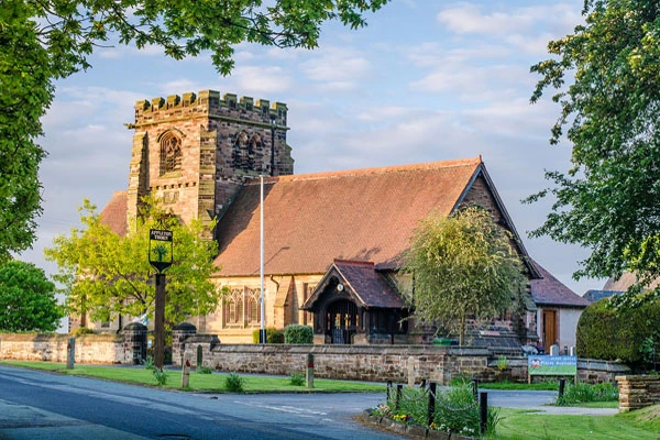 Stone church with crenellated tower, terracotta roof, and wooden porch, surrounded by trees and green churchyard
