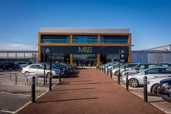 M&S store exterior with orange and dark facade, parked cars in foreground, clear blue sky