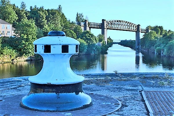 Blue cylindrical structure on riverbank with metal bridge and trees in background