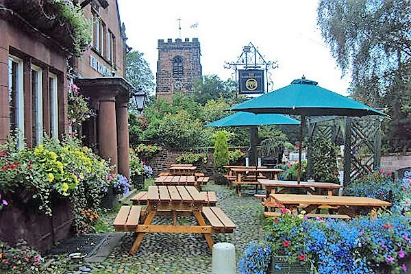 Outdoor seating area with wooden picnic tables under a turquoise umbrella, stone church tower visible in background