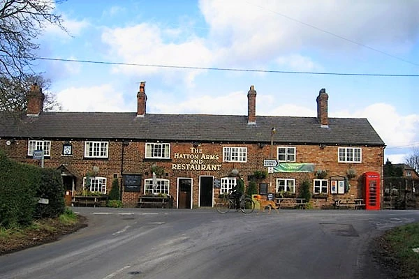 The Hatton Arms pub, a brick building with multiple windows and chimneys, viewed from the road with a red telephone box to the right