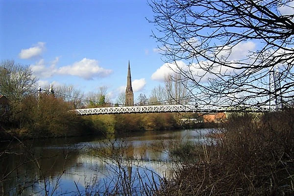 River with bare trees, metal bridge, and church spire in background under blue sky