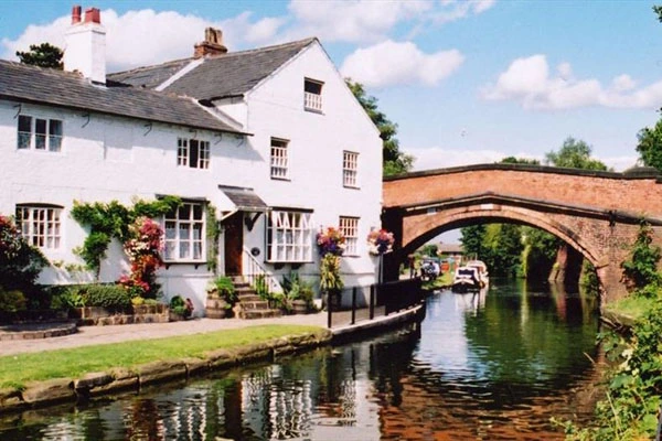 White cottage beside a canal with brick bridge, flowering plants by the entrance, and moored boat visible under the arch