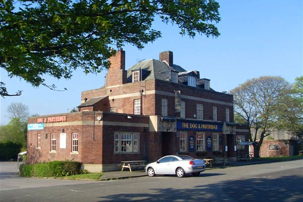 Red brick pub building with "The Dog & Partridge" signage, white car parked outside, trees and clear sky