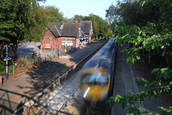 Train passing a brick railway station building with platforms and surrounding trees