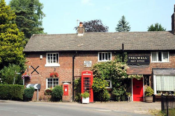 Red-brick cottage with slate roof, featuring two red post boxes and a red door, with ivy and climbing plants, and a "Thelwall Post Office" sign visible on the right side