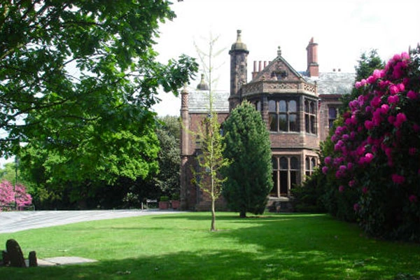 Victorian mansion with red brick and stone detailing, viewed across a manicured lawn with flowering shrubs and mature trees