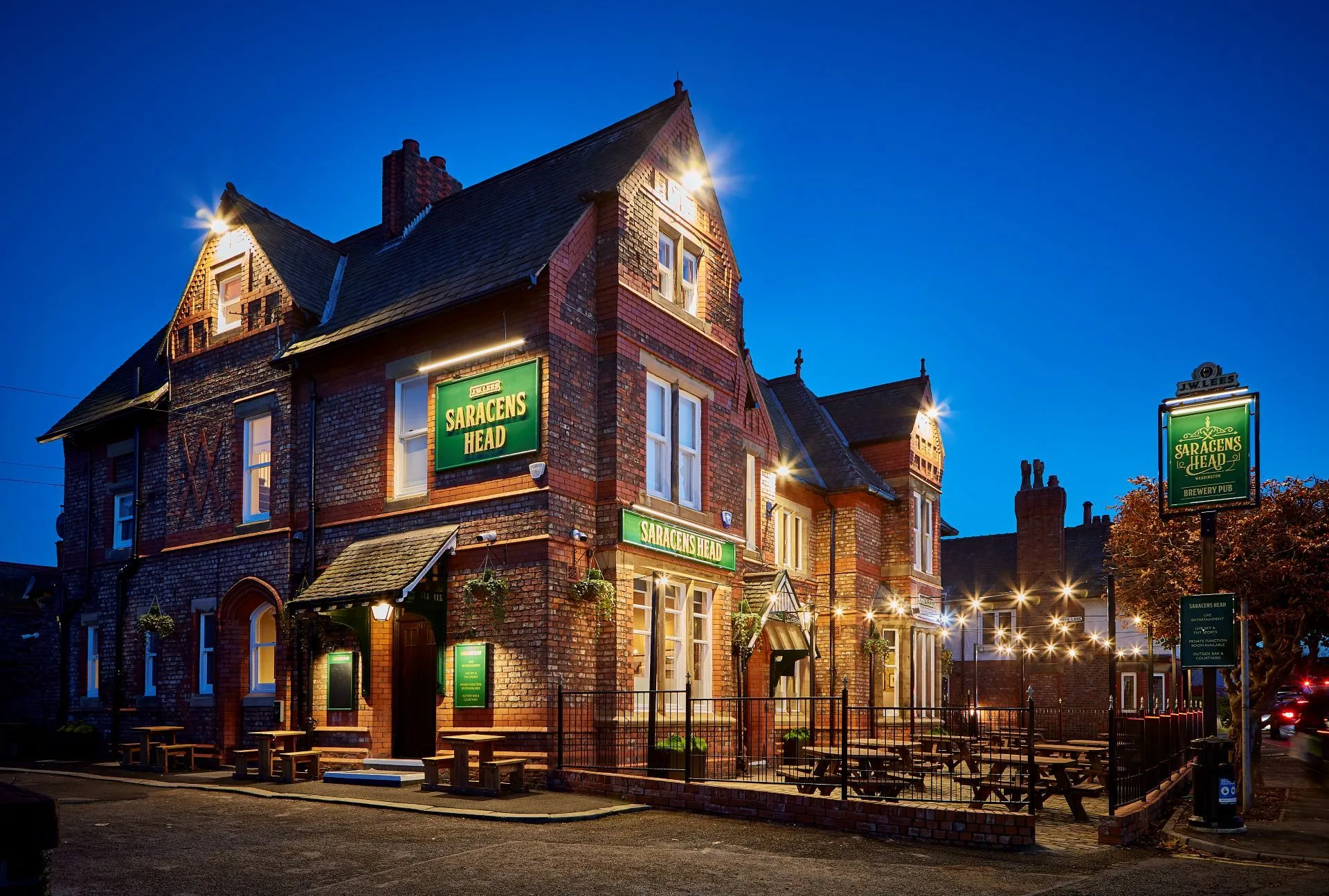 Red brick pub building lit up at dusk with green "Saracens Head" signs, metal railings, and benches outside