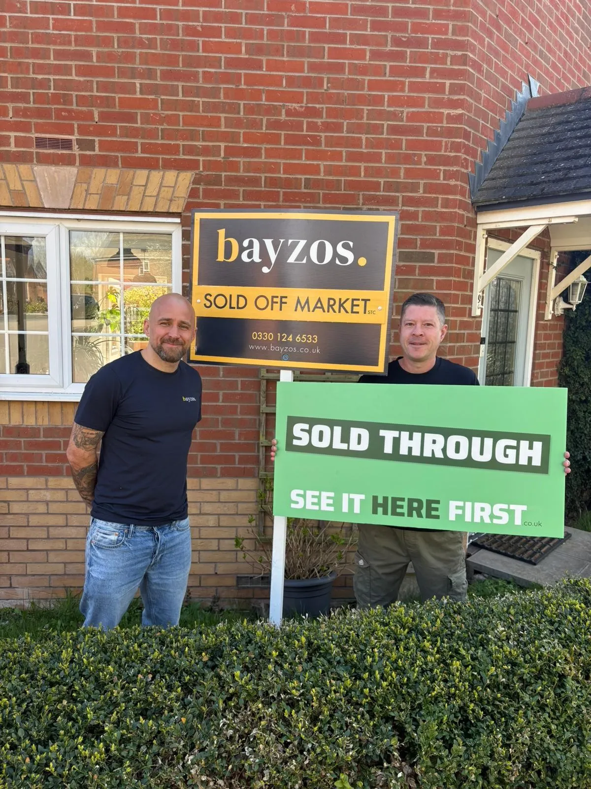 Gary, the founder of Bayzos, stands with a happy homeowner. They stand outside a house next to a sold off market board. The owner is holding a sign saying "sold through see it here first"