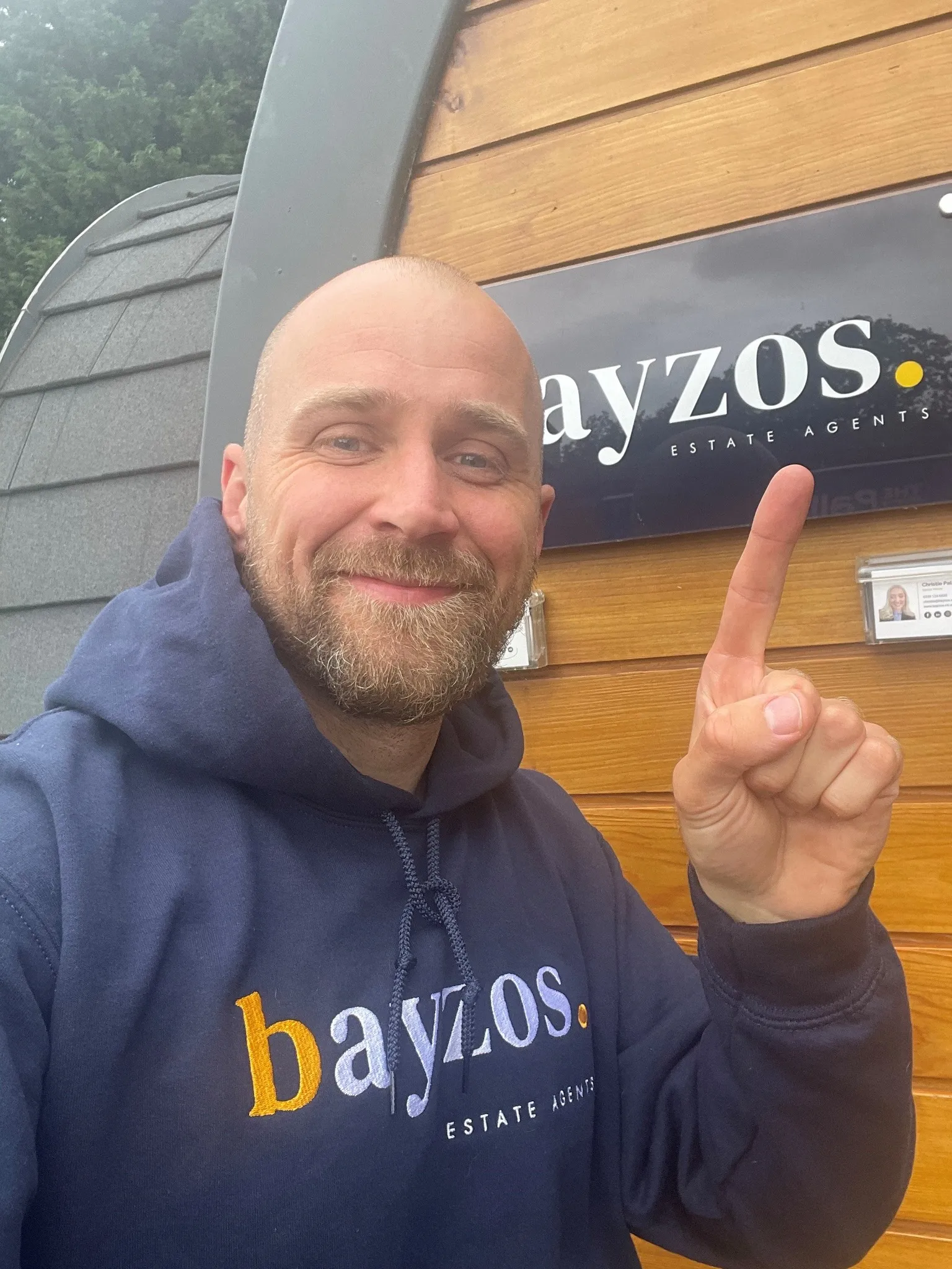 Gary, the founder of Bayzos, is wearing a Bayzos branded hoody and pointing at a Bayzos Estate Agents branded sign