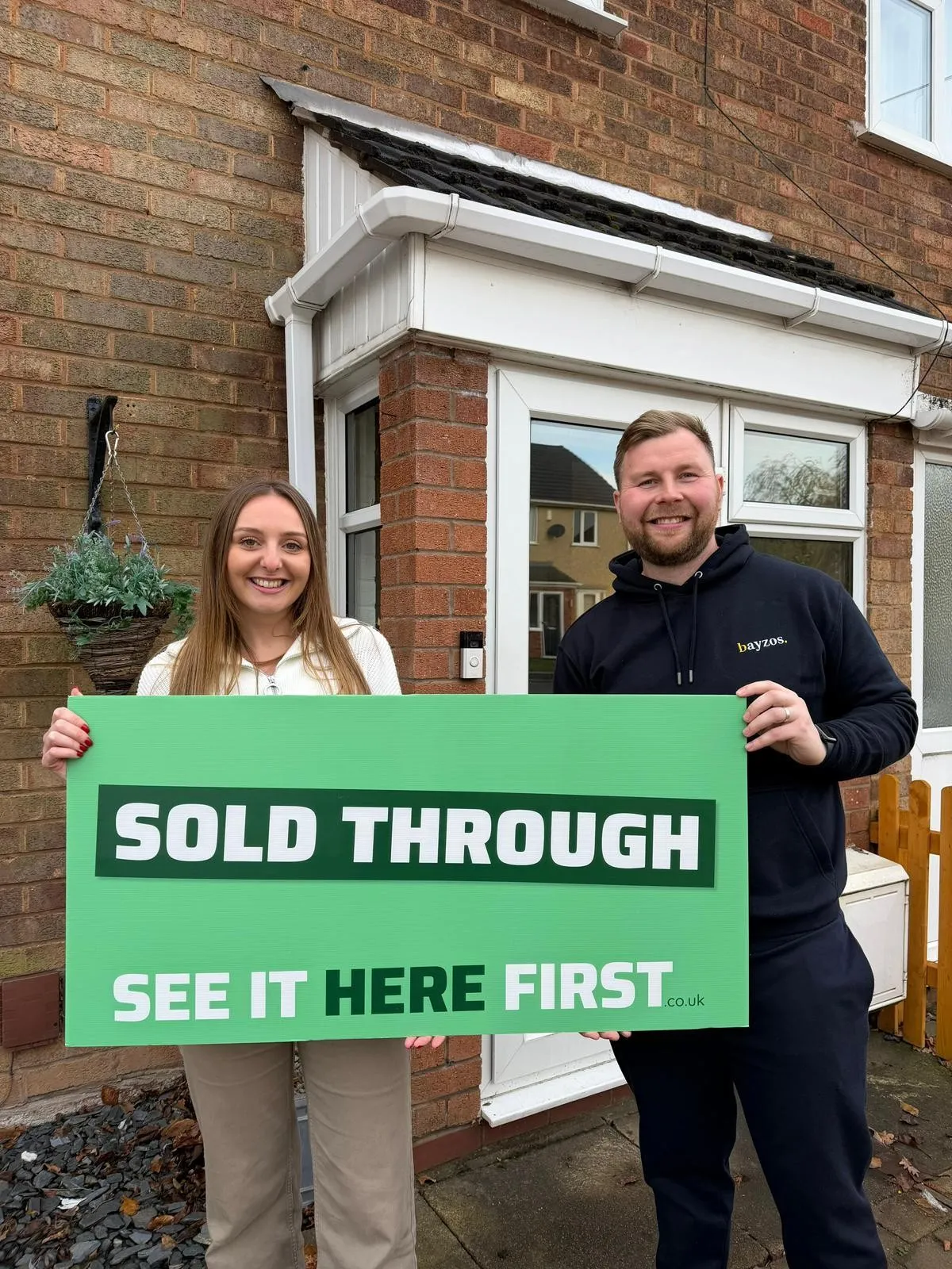 James from Bayzos Estate Agents stands outside a house with a smiling homeowner. They're both holding a board saying "Sold Through See it Here First"