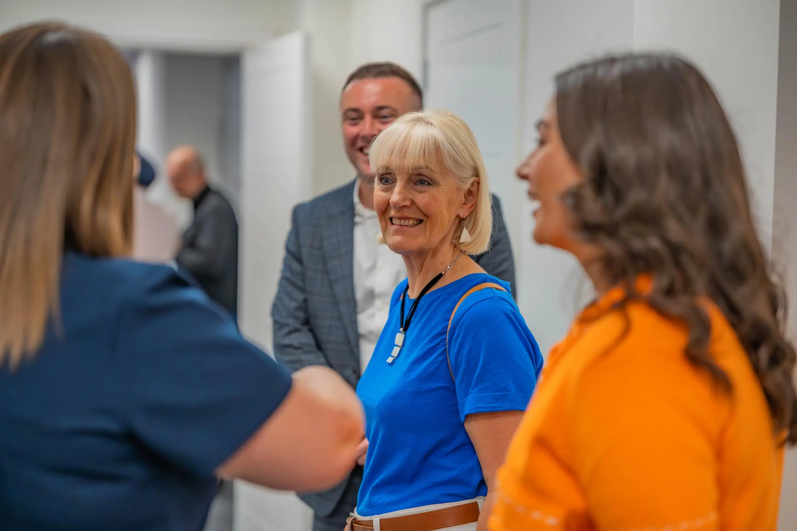 Four people conversing indoors in a white-walled space. Woman in blue shirt with lanyard in center, smiling.