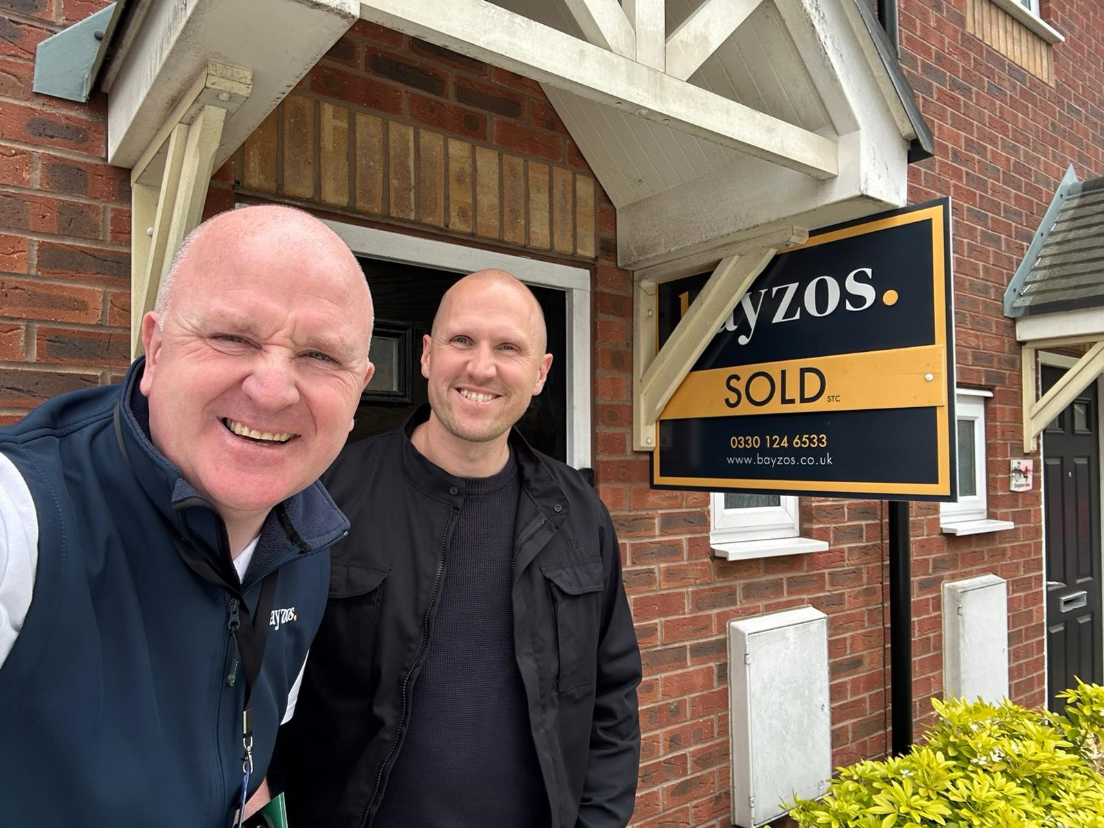 Phill, an estate agent for Bayzos, stands next to a house with a sold board outside and a smiling homeowner