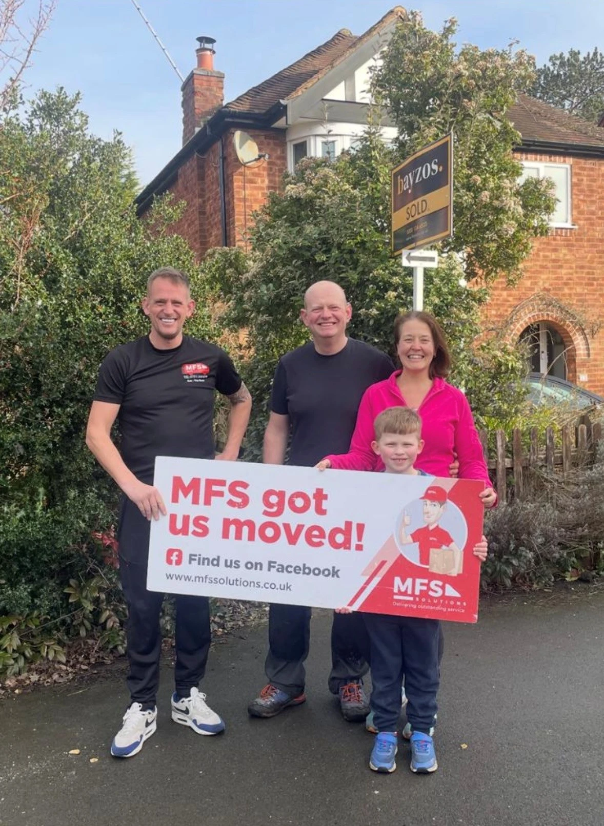 Four people standing outside a brick house holding a sign reading "MFS got us moved!" with a Bayzos "SOLD" sign visible behind them