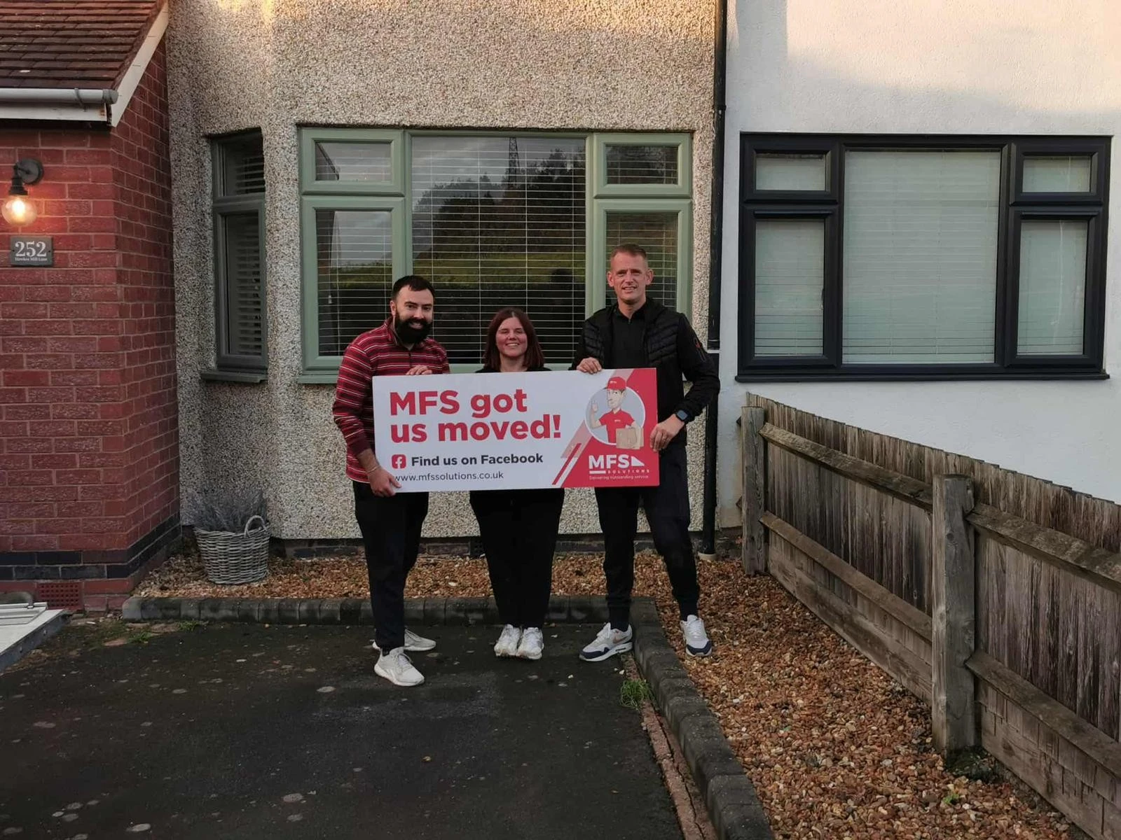 Three people holding a red banner reading "MFS got us moved!" outside a house with grey render and green-framed windows, house number 252 visible