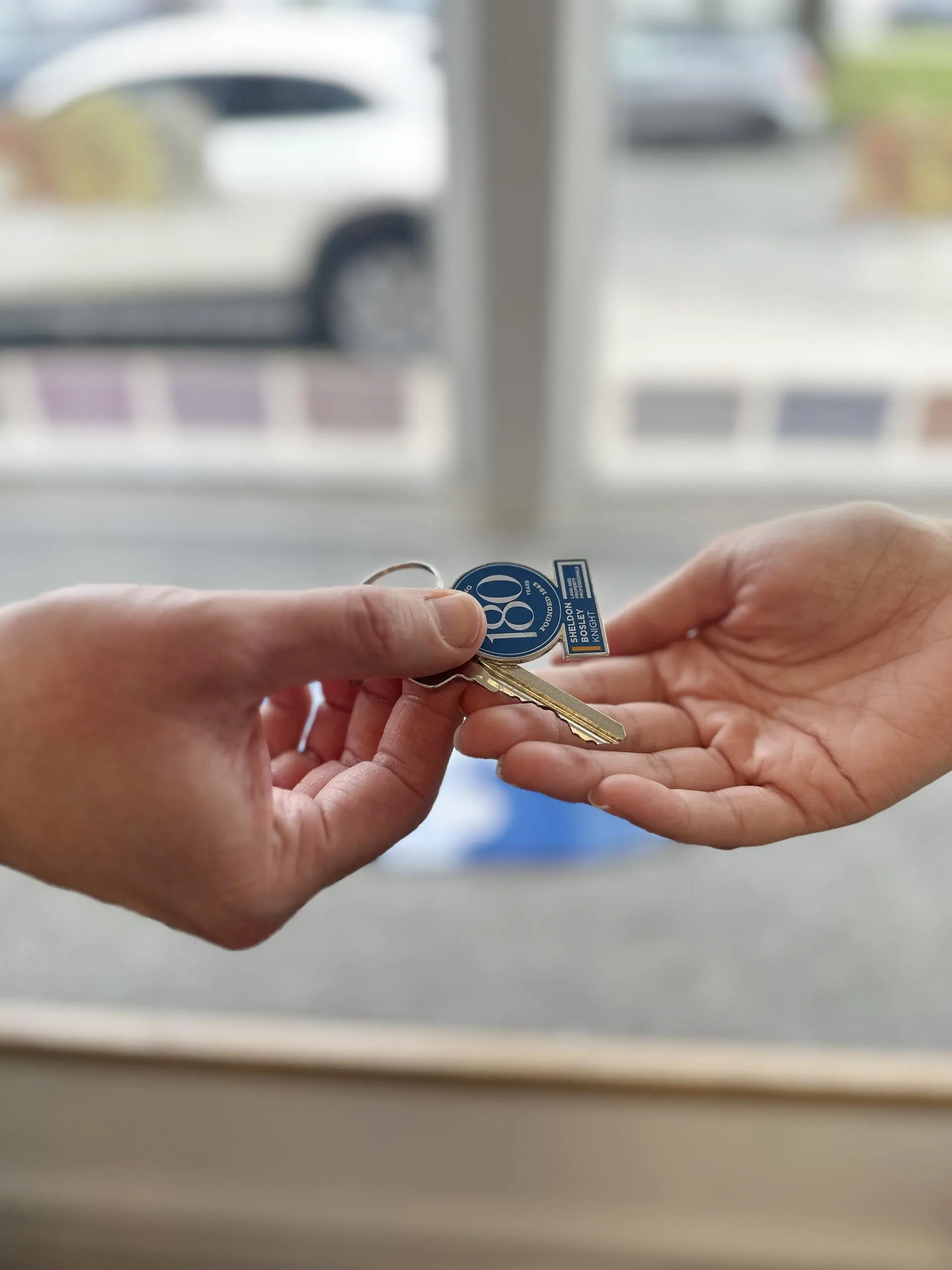 Two hands exchanging a house key with a blue circular keyring, parked cars visible in the background