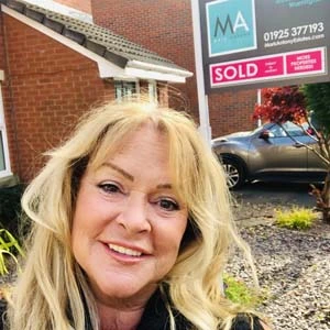 Woman with blonde wavy hair smiling at camera, standing in front of a brick house with a sold sign