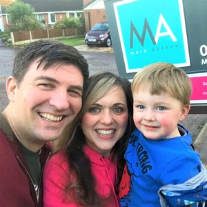 Three people smiling at camera with sold board and house in background