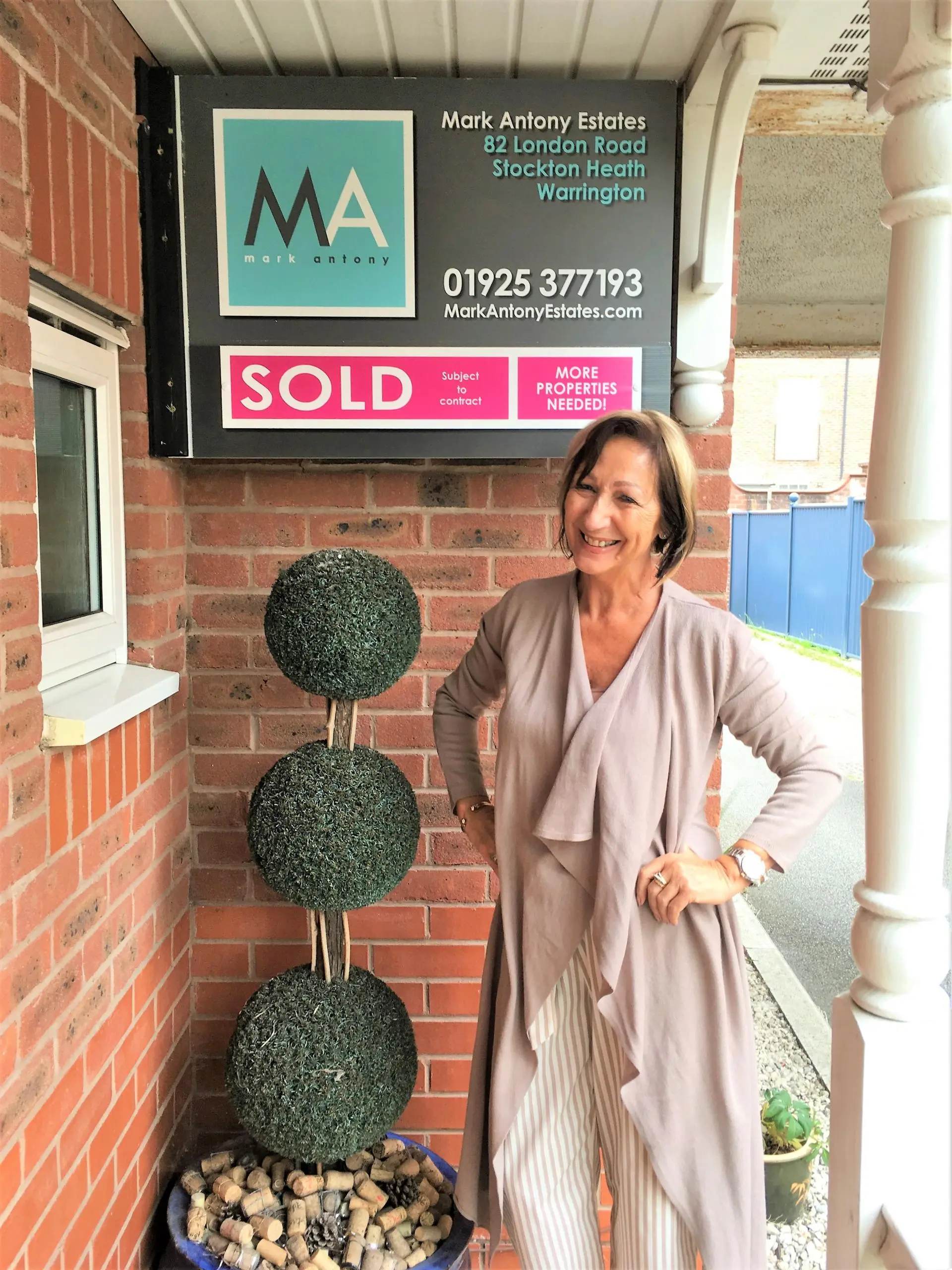 Woman standing outside brick building next to topiary plants, with Mark Antony Estates "SOLD" sign above
