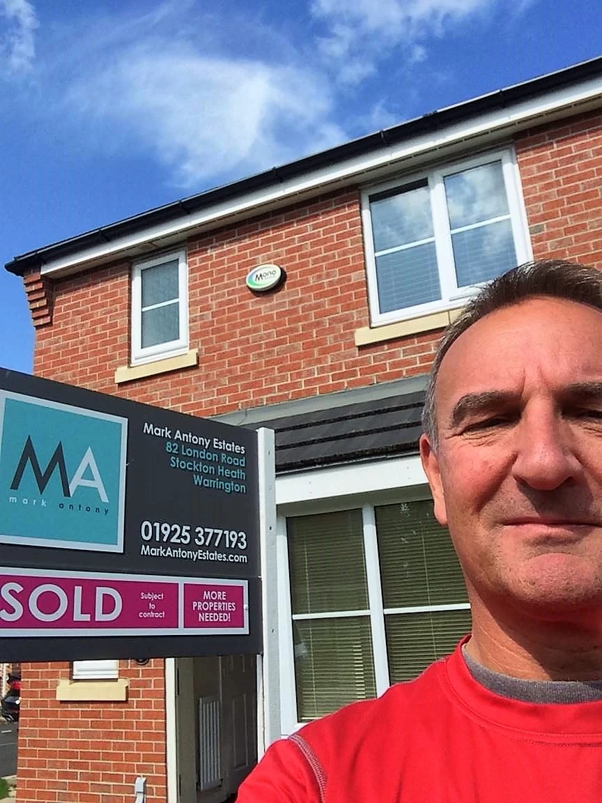 Man in red shirt standing in front of brick building with "SOLD" estate agent sign for Mark Antony Estates