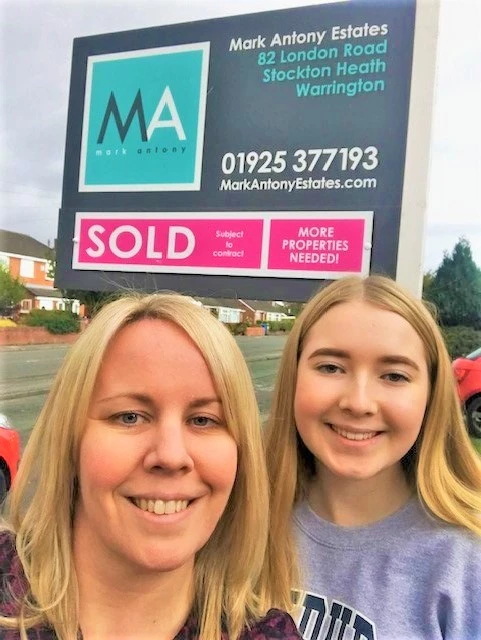 Two people smiling in front of a Mark Antony Estates property board displaying "SOLD" and "MORE PROPERTIES NEEDED!"