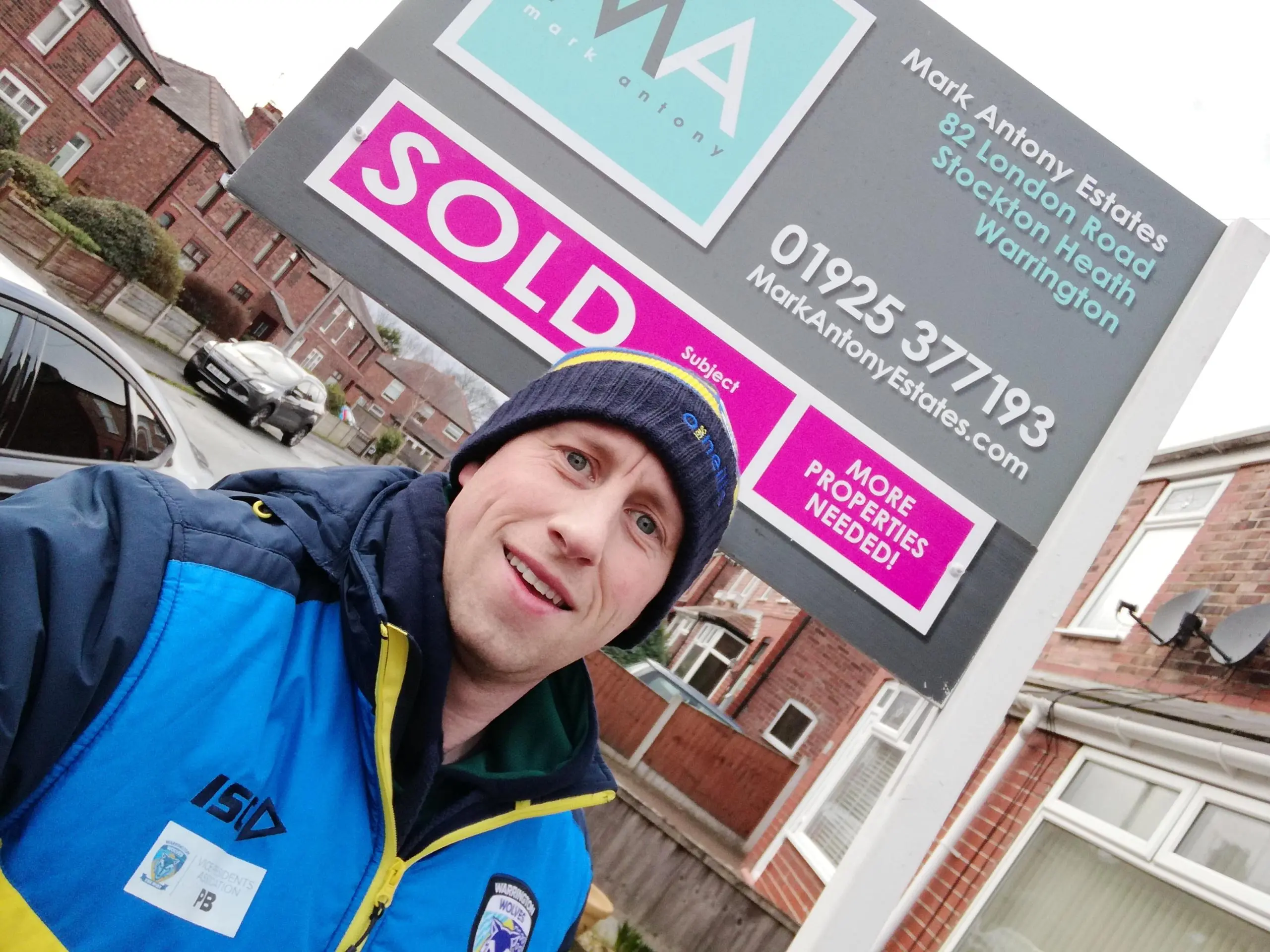 Person in blue and navy jacket and navy beanie smiling at camera next to estate agent "Sold" board displaying Mark Antony Estates details and "More Properties Needed" text