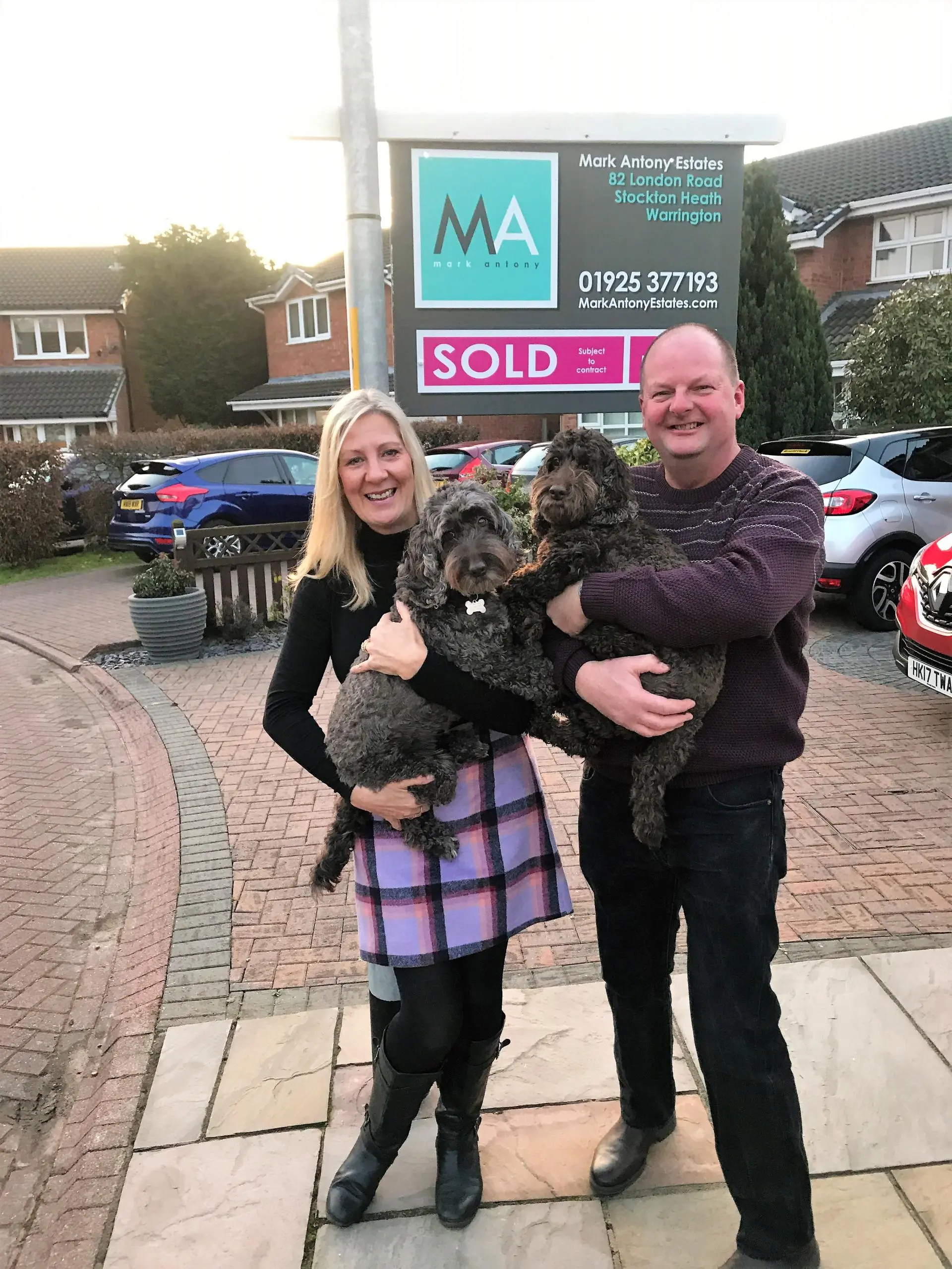 Two people holding two dark dogs in front of a "SOLD" estate agent sign for Mark Antony Estates, Stockton Heath, Warrington