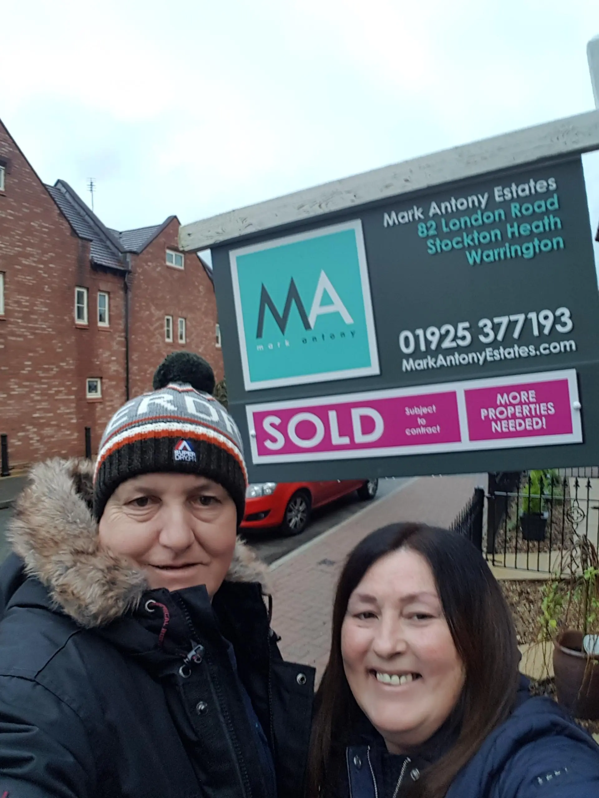 Two people posing in front of a Mark Antony Estates property board displaying "SOLD" and "MORE PROPERTIES NEEDED" with a brick building behind