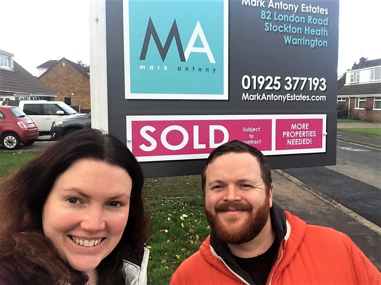 Two people smiling at camera in front of a Mark Antony Estates property board displaying "SOLD Subject to contract" and "MORE PROPERTIES NEEDED!"