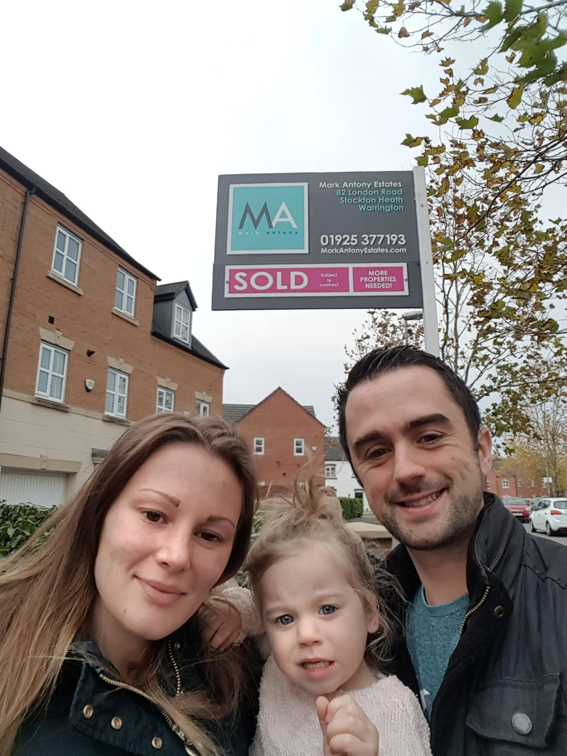 Three people standing in front of a brick building with a Mark Antony Estates "SOLD" sign above them