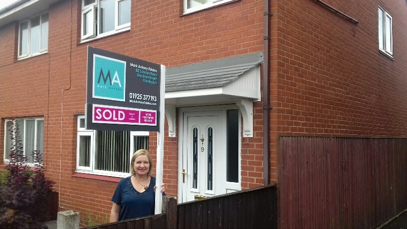 Woman standing outside a red brick house with a "SOLD" estate agent board above the door