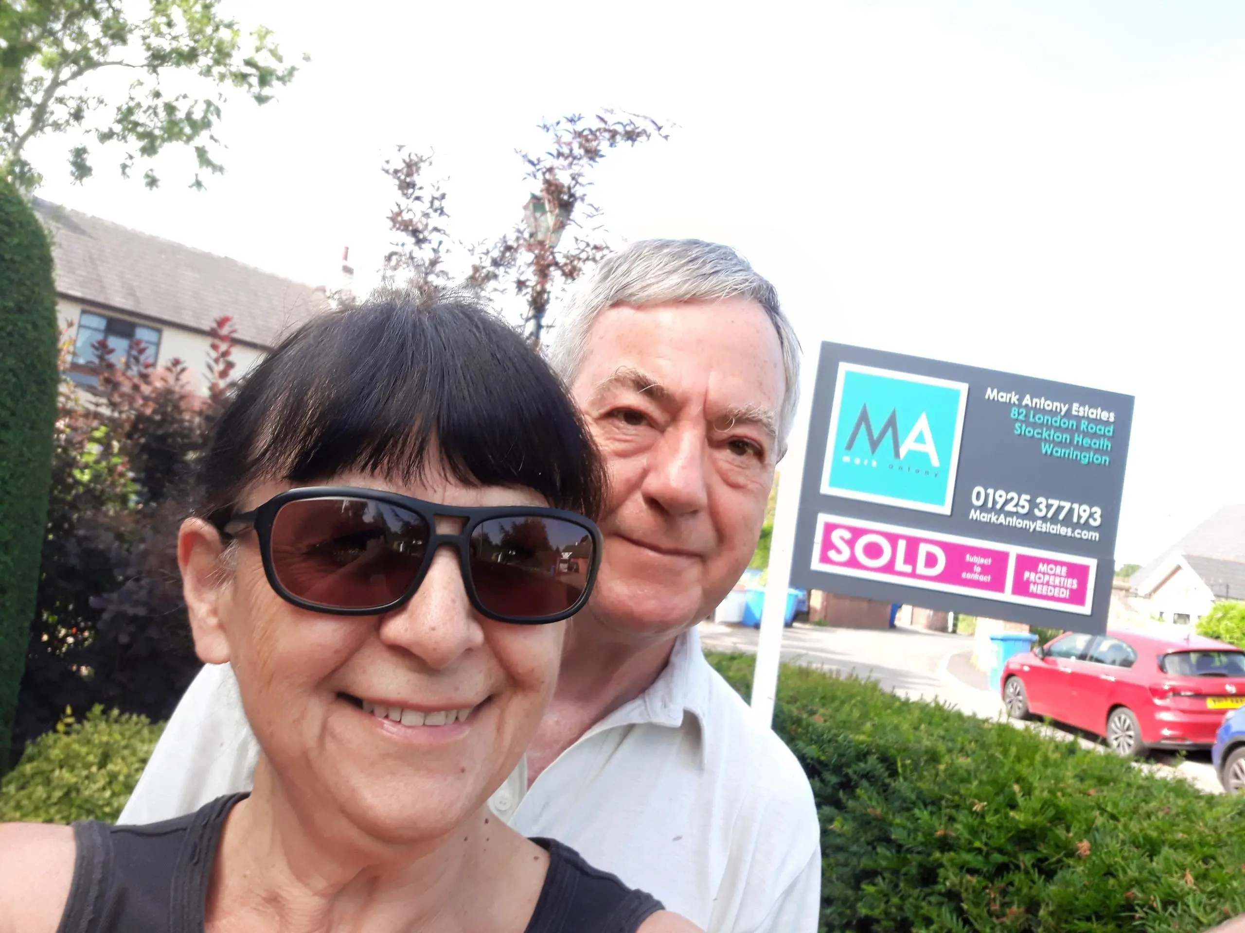 Two people smiling at camera with a "SOLD" estate agent board behind them