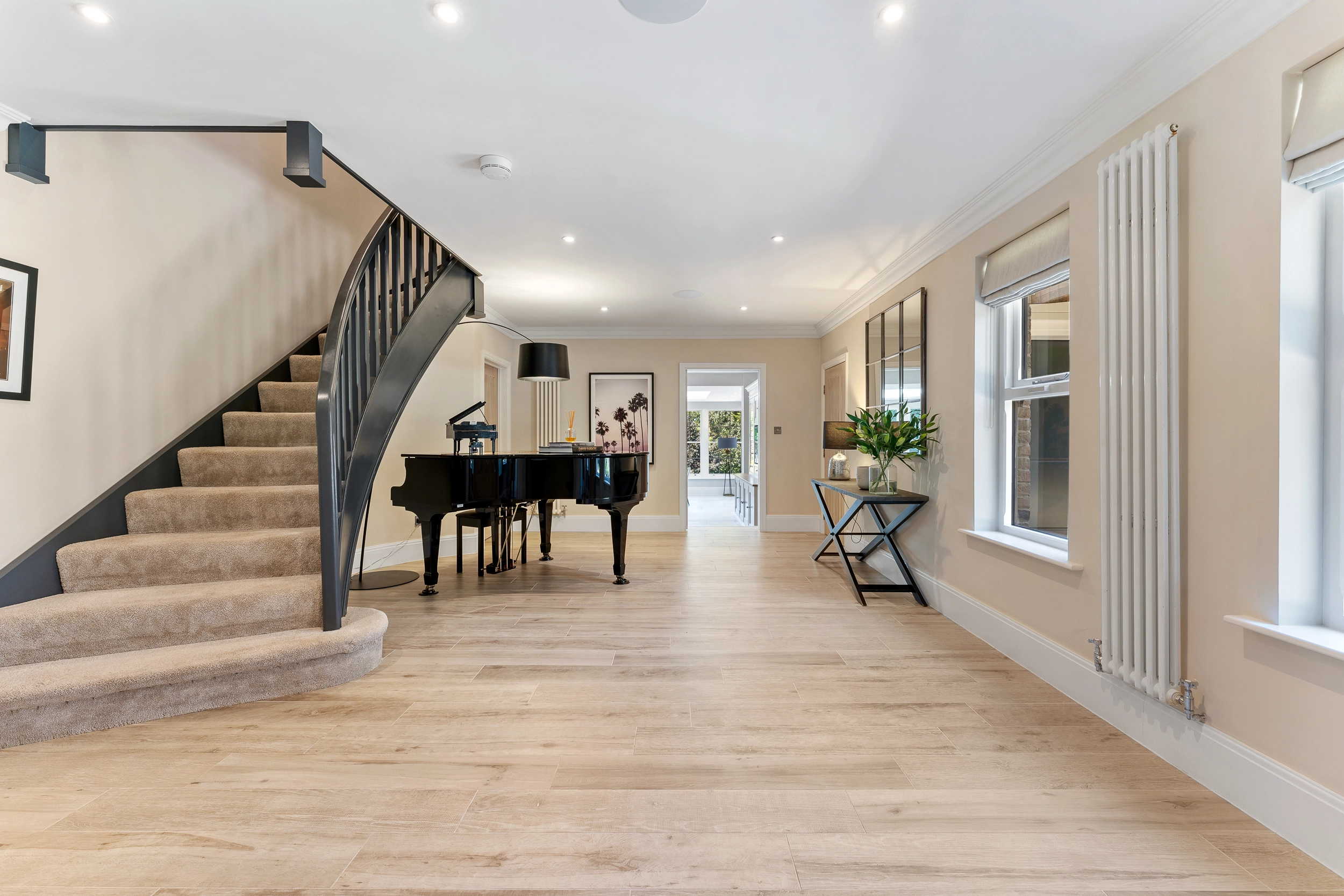 Spacious open-plan hallway with curved staircase, black grand piano, wooden flooring, cream walls, and large windows with curtains