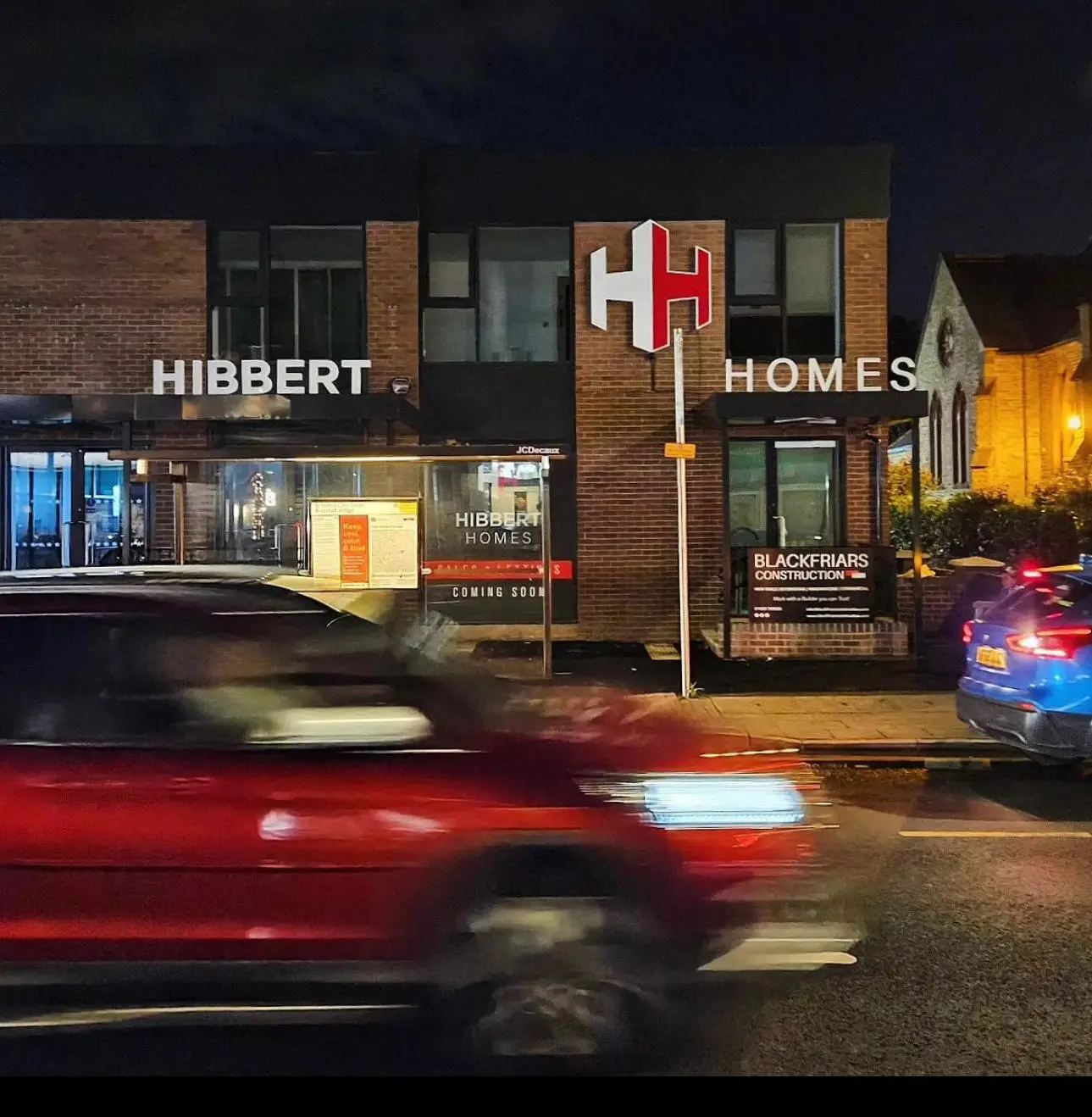 Hibbert Homes storefront at night with illuminated signage, brick facade, and display windows showing 'Coming Soon' and Blackfriars Construction branding