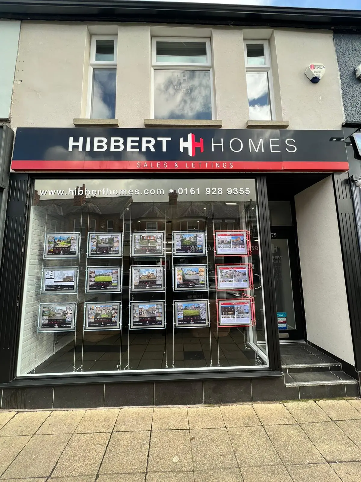 Hibbert Homes estate agency storefront with sales and lettings signage, website www.hibberthomes.com and phone number 0161 928 9355, property listings displayed in window