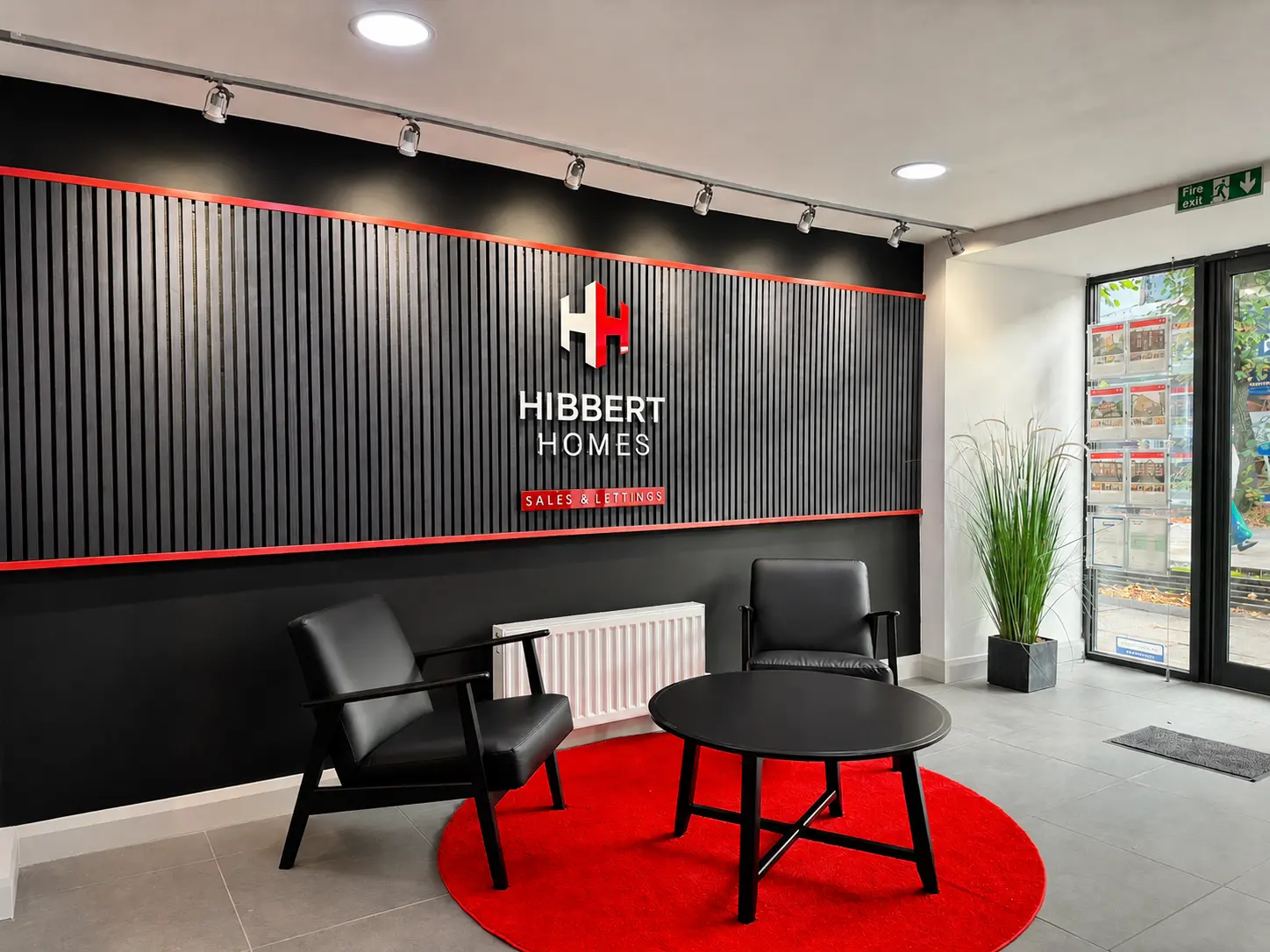 Hibbert Homes estate agency office interior with black and red branding on corrugated wall, black leather chairs, round black table on red circular rug, and property listings visible through glass door