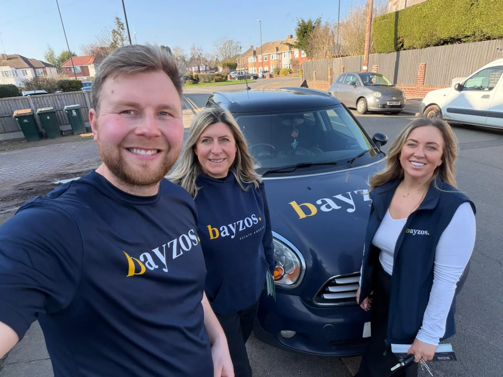 James, Becky, and Leah from Bayzos Estate Agents wear bayzos branded tops and stand in front of a bayzos branded car