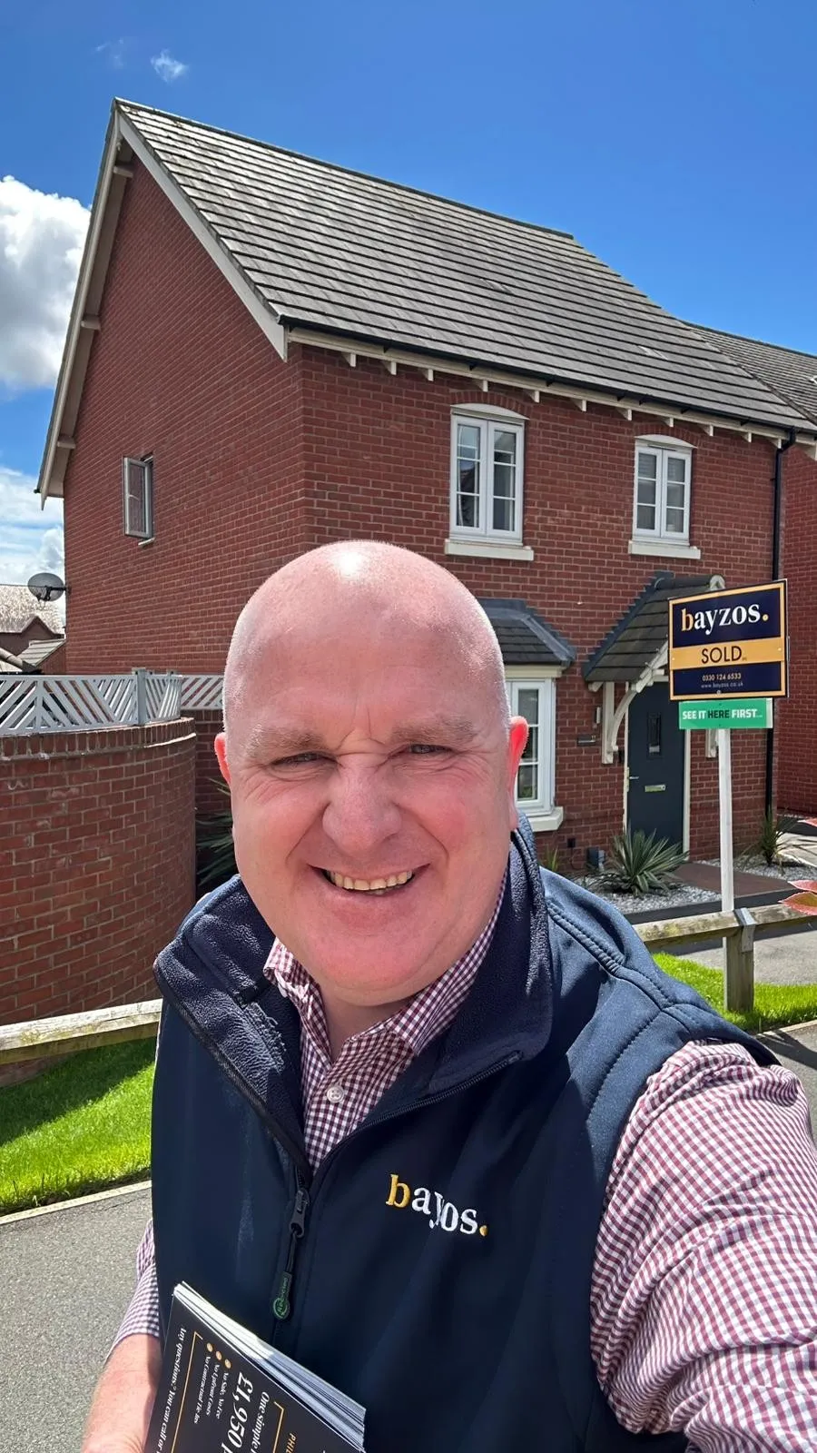 Man in navy Bayzos gilet standing in front of red brick house with "SOLD" sign