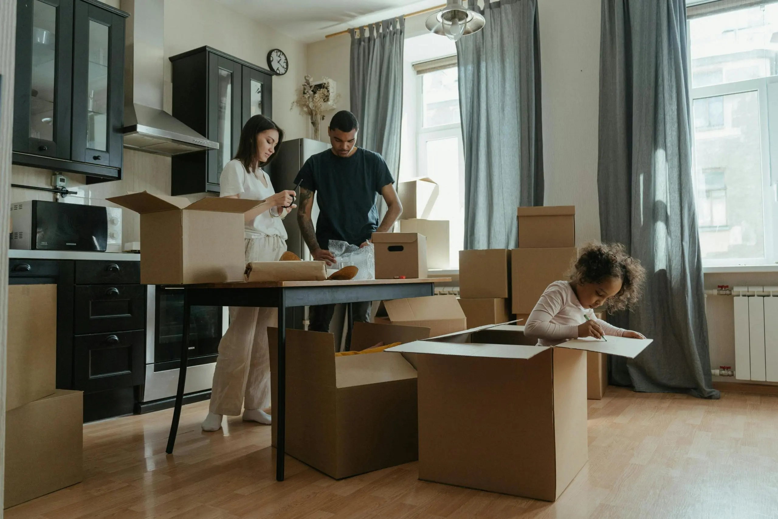 Modern kitchen with black cabinetry, two adults and a child unpacking cardboard boxes, grey curtains and wooden flooring