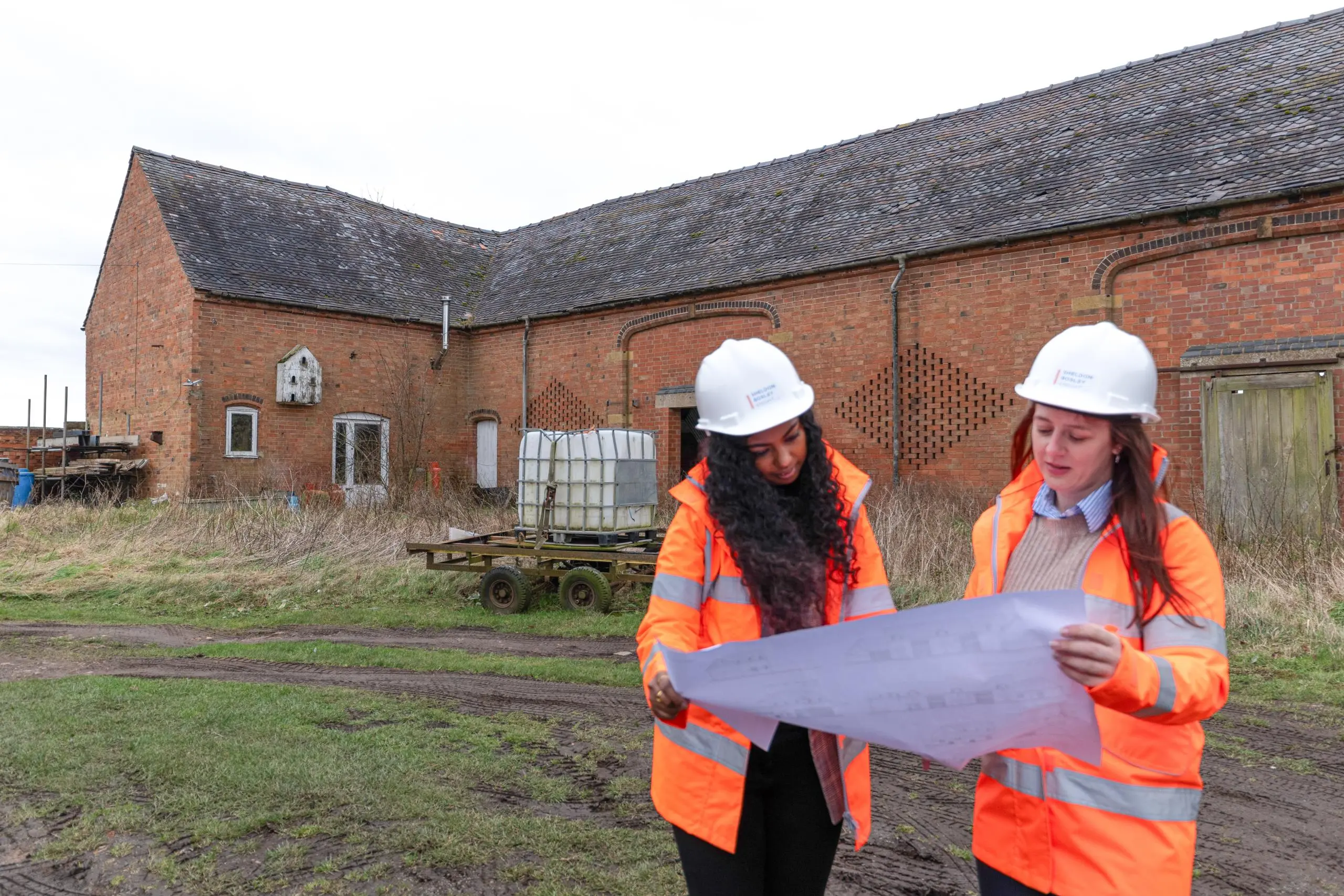 A planning team stand looking at architectural plans in front of a rural outbuilding