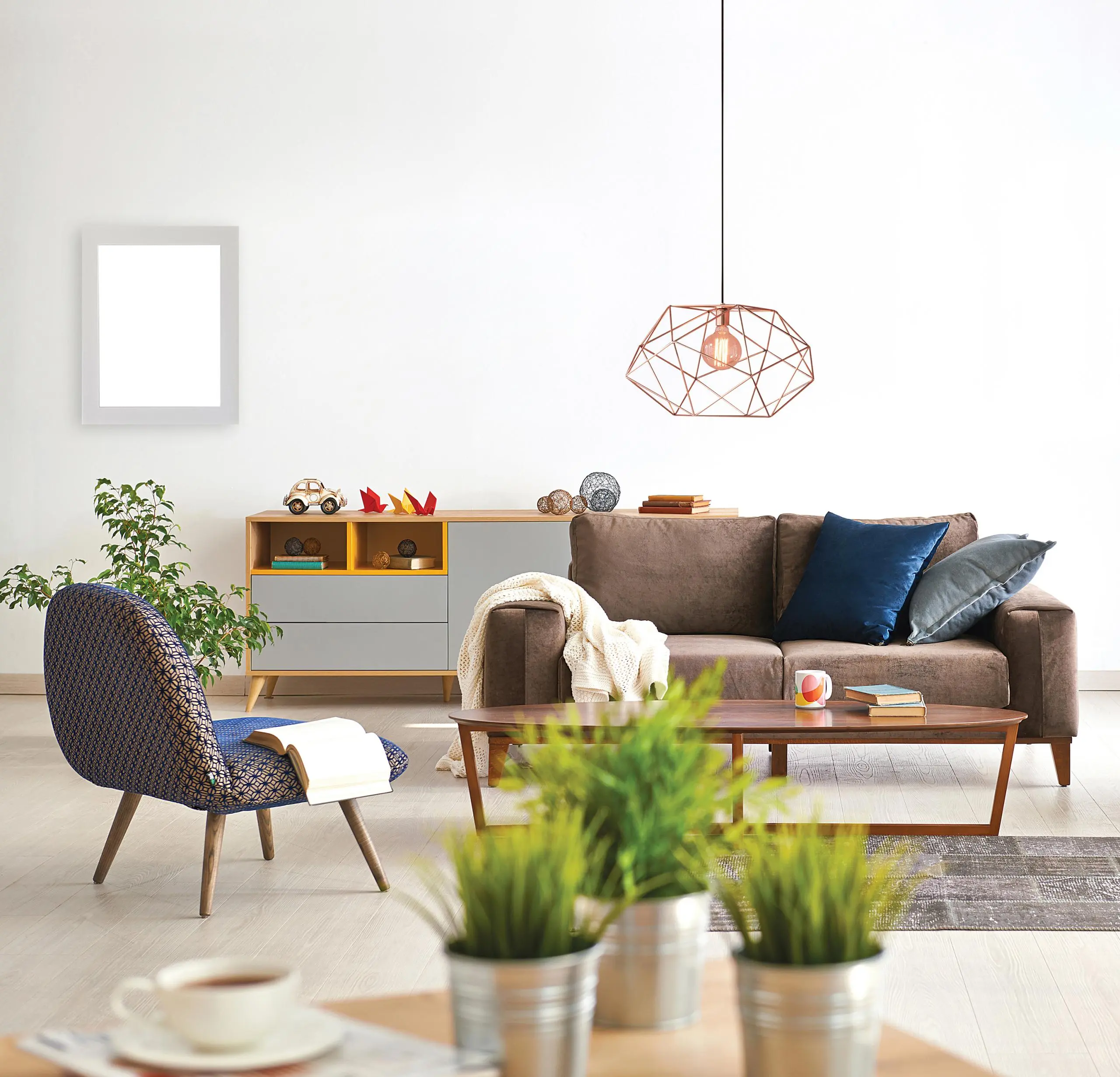 Modern living room with brown sofa, black woven chair, wooden coffee table, geometric copper pendant light, storage unit with shelves, potted plants, and framed artwork on white wall