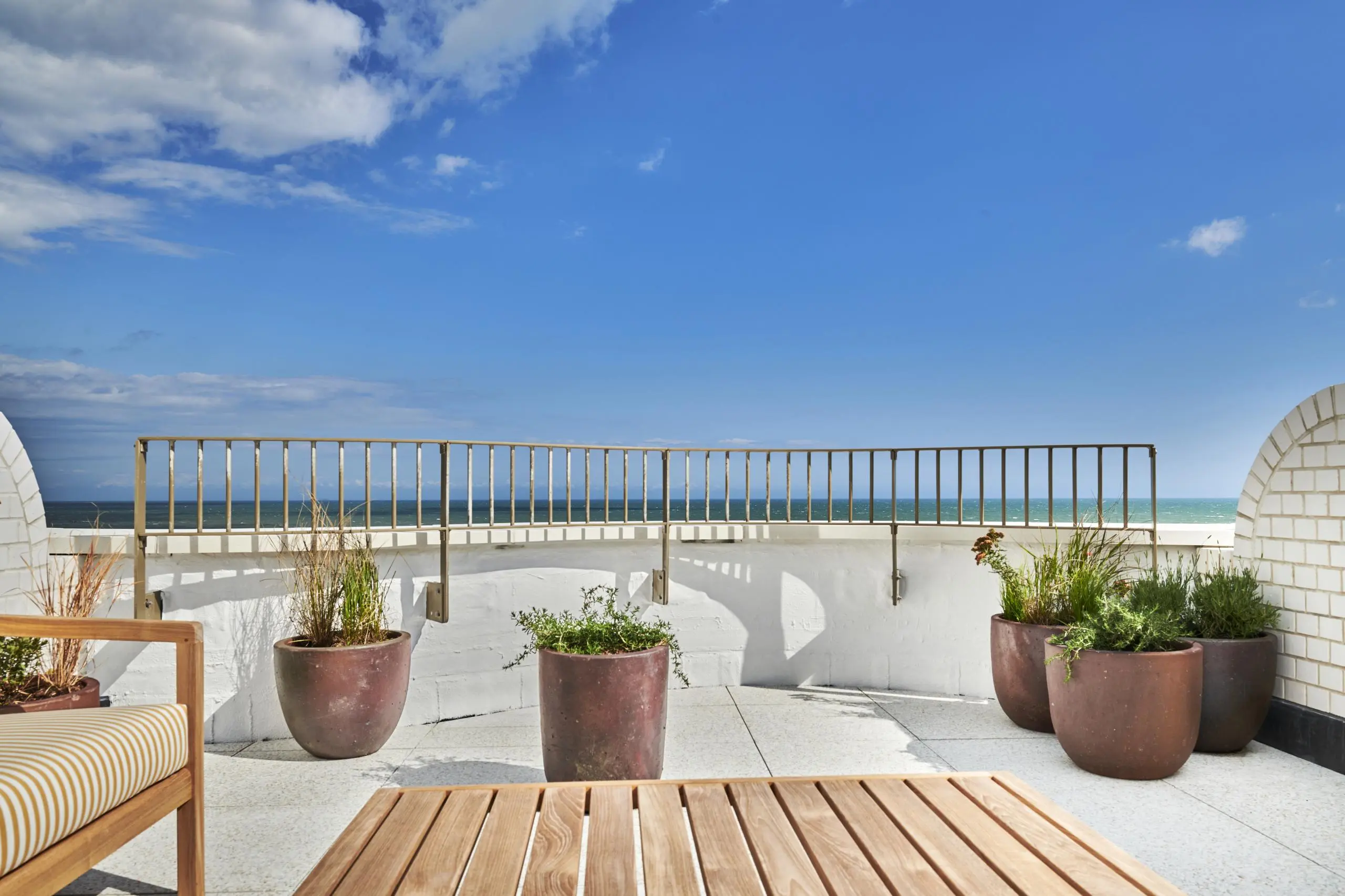 Rooftop terrace with sea view, metal railings, wooden decking, terracotta planters with greenery, and white brick walls