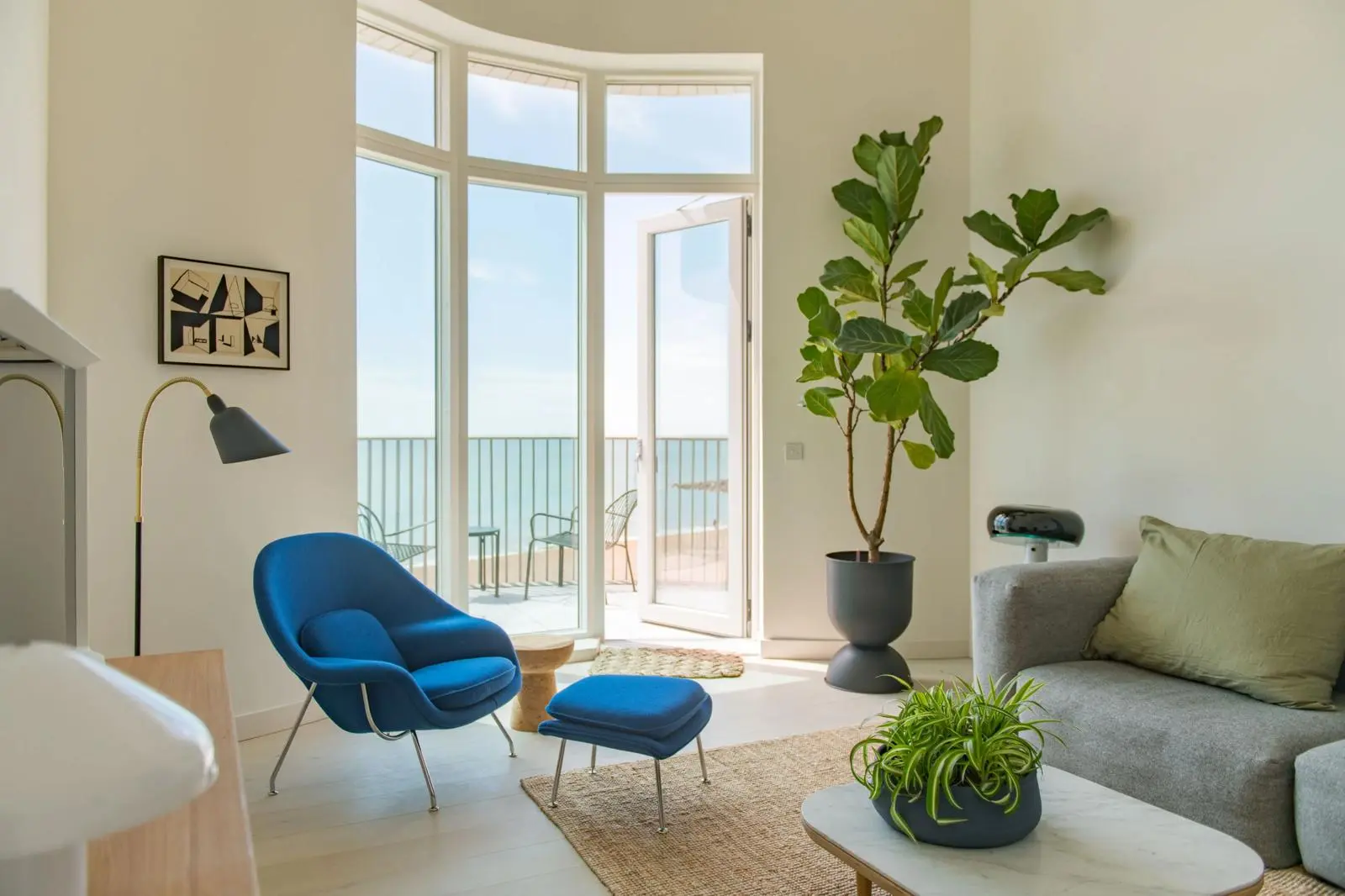Modern living room with blue armchair and footstool, grey sofa, potted plants, arc floor lamp, and floor-to-ceiling glass doors opening onto a balcony with sea view