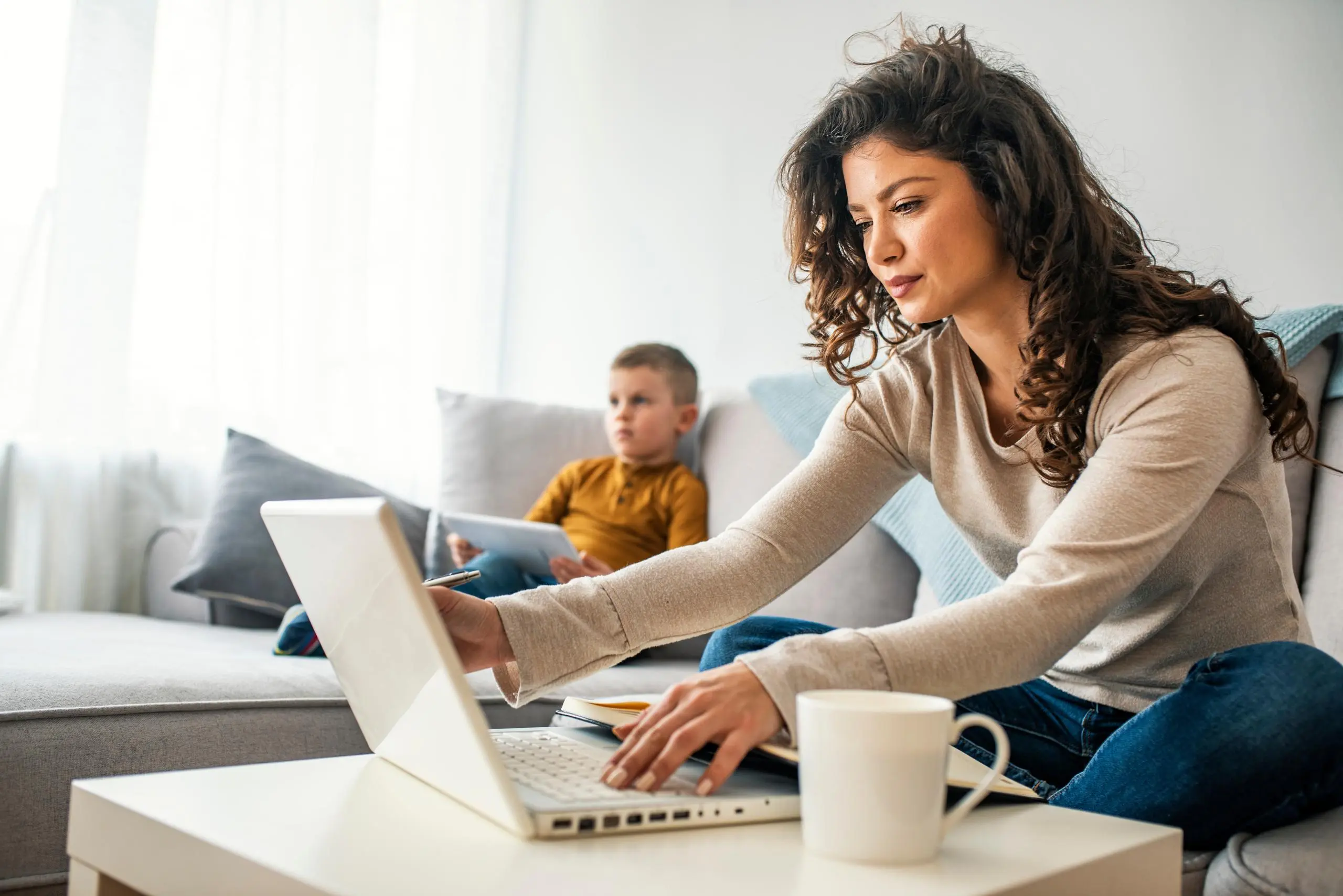 Woman using laptop on sofa with child in background